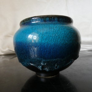 Peut inclure: Un bol en céramique rond présentant un dégradé vibrant de glaçure turquoise et bleu profond avec une finition craquelée prononcée. La partie supérieure affiche une teinte cyan vive qui passe au bleu marine foncé, finissant sur une base texturée.