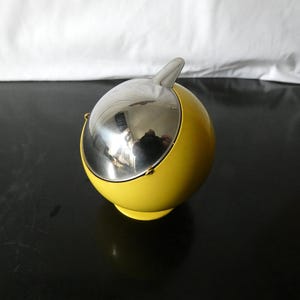 Può includere: Un posacenere sferico giallo con un coperchio argentato incernierato e una piccola maniglia. Il posacenere ha una base circolare e una finitura liscia e lucida. Il coperchio è riflettente.