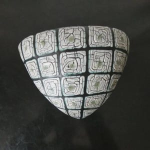 Può includere: Una ciotola in ceramica triangolare caratterizzata da un motivo a griglia con linee nere che separano quadrati bianchi. Ogni quadrato presenta un design a spirale concentrica in rilievo con una texture crepata e sfumature verde chiaro.