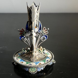 Op de afbeelding: Een sierlijke zilverkleurige metalen kandelaar met kleurrijke emaille accenten. De basis is rond met blauw, groen en wit emaille, en de centrale stam heeft ingewikkelde filigraan- en bloemmotieven. De houder is ongeveer 15 cm hoog.