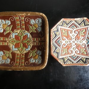 Könnte beinhalten: Zwei dekorative Keramikfliesen mit erhabenen Reliefmustern. Die quadratische Fliese hat einen tiefroten Hintergrund mit hellblauen und grünen Blumenmotiven. Die achteckige Fliese zeigt ein geometrisches Design in Rot, Grün, Schwarz und Weiß.