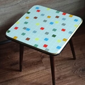 Peut inclure: Une table d'appoint carrée avec un plateau blanc orné d'un motif en grille composé de petits carrés multicolores rouges, bleus, jaunes, verts et gris. La table possède un bord métallique fin et quatre pieds fuselés en bois brun foncé.
