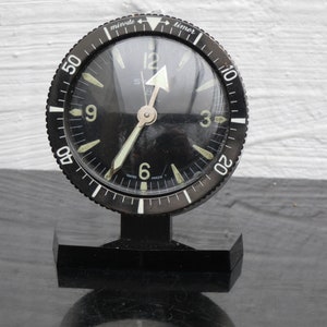 Op de afbeelding: Een vintage zwarte en zilveren timerklok met een ronde wijzerplaat. De klok heeft een zwarte lunette met witte minutenmarkeringen en een draaiende timerwijzerplaat. De wijzerplaat toont de cijfers 3, 6, 9 en 12 in een lichte kleur, met een zilveren wijzer.