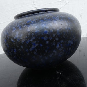 Può includere: Un vaso di ceramica blu scuro con un motivo a pois blu. Il vaso ha un corpo arrotondato e un bordo leggermente svasato. La superficie ha una finitura lucida, che riflette la luce.