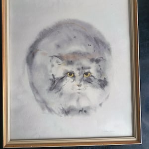 Puede incluir: Pintura a la acuarela de un gato con ojos amarillos, enmarcada en un marco dorado. El pelaje del gato está representado en tonos grises y blancos, creando una apariencia suave y esponjosa. La obra de arte está sobre un fondo claro.
