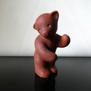 Può includere: Una piccola statuetta di orso in piedi realizzata in un materiale opaco color marrone rossiccio. L'orso ha una testa arrotondata con due piccole orecchie, semplici rientranze per gli occhi e un muso rialzato. Un braccio è teso verso l'esterno.