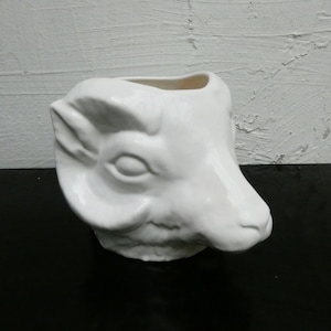 Può includere: Una fioriera a forma di testa di ariete in ceramica bianca. La fioriera presenta dettagli del viso, tra cui occhi, orecchie e muso. L'interno è cavo, progettato per contenere piante o altri oggetti decorativi. Su sfondo bianco e nero.