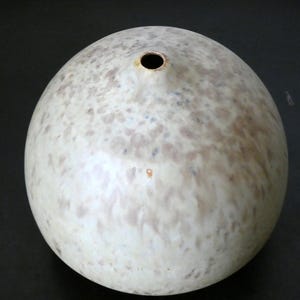 Può includere: Un vaso in ceramica sferico con una liscia smaltatura color bianco sporco caratterizzata da macchie screziate marrone chiaro e grigie su tutta la superficie. Il vaso presenta una piccola apertura circolare al centro, creando una forma arrotondata.