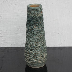 Può includere: Un vaso in ceramica alto e affusolato caratterizzato da una superficie ruvida e fortemente strutturata in tonalità verde acqua profondo e verde spento. L'esterno presenta un motivo in rilievo simile a una rete con sfumature marroni, mentre il bordo interno rivela una finitura in argilla liscia.