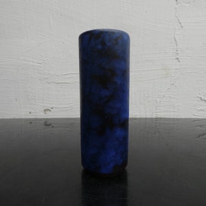 Può includere: Un vaso cilindrico con un motivo screziato blu scuro e nero. Il vaso è alto e sottile, con una parte superiore liscia e arrotondata e una base scura. Lo sfondo è un muro bianco strutturato e una superficie nera.