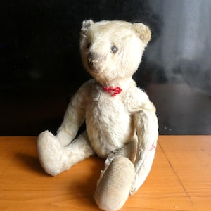 Può includere: Un orsacchiotto vintage color crema, seduto su una superficie di legno. L'orso ha un fiocco rosso intorno al collo e un naso cucito. Le sue braccia e gambe sono leggermente usurate, conferendogli un aspetto vissuto.
