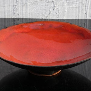 Può includere: Una ciotola poco profonda fatta a mano, con un interno rosso vibrante e un esterno nero. La ciotola ha un bordo ampio e leggermente irregolare e poggia su una piccola base scura. Lo sfondo presenta una superficie strutturata bianca e grigia.