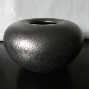 Peut inclure: Un vase rond en céramique gris foncé avec une surface texturée et une large ouverture. Le vase a un éclat métallique, reflétant la lumière. La forme générale est une sphère aplatie, adaptée à la décoration intérieure.