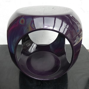 May include: A glossy, deep purple stool with a rounded, cube-like shape featuring large, circular cutouts on each side. The surface has a smooth, reflective finish that catches light, creating a dark, polished appearance with subtle color variations.