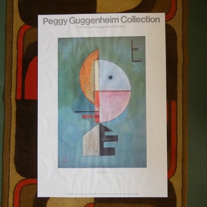 May include: A Peggy Guggenheim Collection poster featuring an abstract geometric design. The artwork includes a large circle divided into sections of orange, pink, and blue, with a blue background. The poster is displayed on a white background.