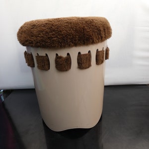 Peut inclure: Un tabouret cylindrique beige avec un dessus en fourrure marron et moelleux. Le tabouret comporte une série d'accents décoratifs en fourrure marron sur la partie supérieure de l'extérieur. Le tabouret est posé sur un fond blanc.