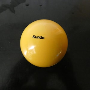 Puede incluir: Una bola amarilla brillante con la palabra "Kundo" impresa en negro. La bola es lisa y reflectante, con un acabado brillante. El objeto es una esfera.