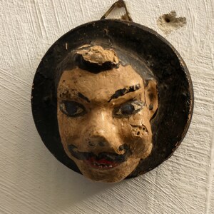 Pode incluir: Uma máscara vintage pintada à mão com um rosto estilizado. A máscara apresenta um rosto bronzeado com cabelo preto, um bigode preto e lábios vermelhos. É montada em uma base circular preta e está pendurada em uma parede branca.