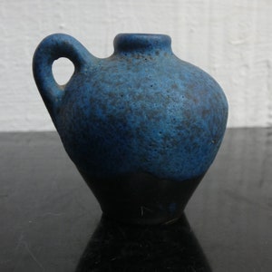 Puede incluir: Un pequeño jarrón de cerámica con asa, con un esmalte azul moteado y texturizado en la parte superior y una base oscura, casi negra. El jarrón tiene un cuerpo redondeado y una pequeña abertura.