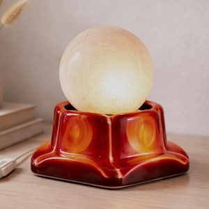 Op de afbeelding: Een retro tafellamp met een bolvormige, crèmekleurige glazen kap. De basis is een glanzend, stervormig ontwerp in rood- en oranje tinten. De lamp is aangesloten en staat op een houten oppervlak.