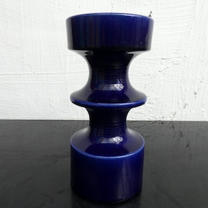 Può includere: Un portacandele in ceramica blu cobalto con una forma a clessidra unica. Il portacandele ha una base e una parte superiore ampie, con una sezione centrale più stretta e scanalata. La finitura lucida riflette la luce, evidenziando le curve e i dettagli.