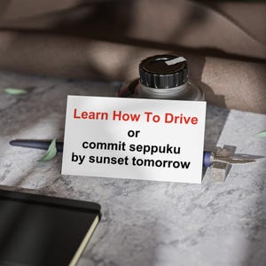 Op de afbeelding: Een stilleven met een witte kaart met de tekst "Learn How To Drive or commit seppuku by sunset tomorrow" in het rood en zwart. Een zwarte inktfles, pen en notitieboek zijn ook zichtbaar.