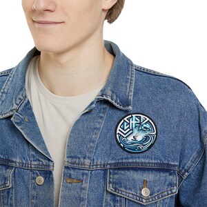 May include: A blue denim jacket with a circular patch featuring a stylized graphic of a person kitesurfing in the ocean. The patch has a white background with blue and black accents.