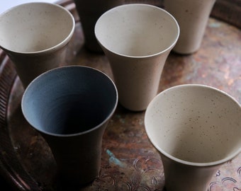 Può includere: Un set di sei tazze in ceramica maculata, in varie tonalità di beige, grigio e blu scuro. Le tazze hanno una forma svasata e sono disposte su un vassoio di metallo decorativo, mostrando un'estetica minimalista.