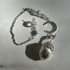 May include: A silver-toned pendulum with celestial charms. The design includes a crescent moon, a sunburst, and an acorn pendant. The chain is adorned with iridescent beads. The pendulum is designed for divination and spiritual practices.