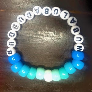 Pode incluir: Um bracelete de contas brancas com a palavra "UBUD" escrita em letras pretas. O bracelete tem contas azuis que desbotam do azul escuro para o azul claro.