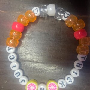 May include: A colourful beaded bracelet with the words "JUICY" and "THICK" spelled out in white beads. The bracelet also features orange, pink, and clear beads, as well as two yellow beads with pink grapefruit slices.