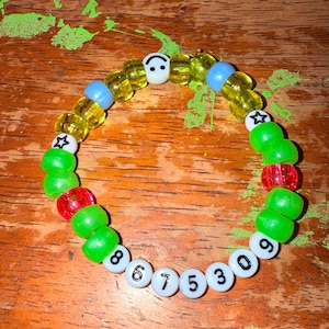 Pode incluir: Um bracelete de contas verdes, amarelas e azuis com um amuleto de carinha sorridente e contas brancas com os números 8675309.