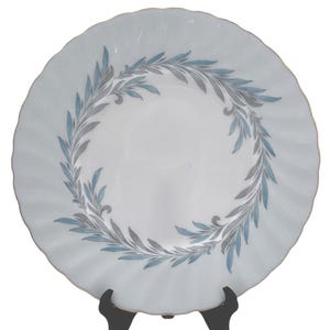 May include: A white ceramic plate with a wavy edge and a gold trim. The plate has a wreath design of blue and grey leaves. The plate is displayed on a black stand, highlighting its decorative pattern.