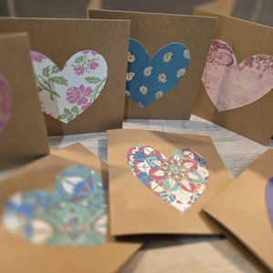 May include: A collection of eight handmade cards with brown paper exteriors and colourful patterned hearts cut out of the centre. The hearts feature various floral, geometric, and abstract designs.