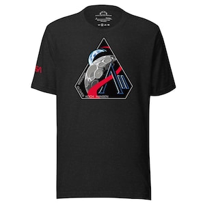 Könnte beinhalten: Dunkelgraues T-Shirt mit einem NASA-Missionsabzeichen mit einem stilisierten Mond, der Erde und roten Akzenten. Die Namen Wiseman, Glover, Koch und Hansen sind auf dem Abzeichen aufgedruckt. Das NASA-Logo ist auf dem linken Ärmel sichtbar.