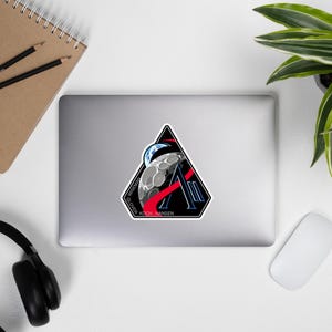 May include: A sticker featuring a triangular design with a black border. The design includes a stylized moon, Earth, and the letter "A". Red and blue accents are present, along with the names "WISEMAN", "GLOVER", "KOCH", and "HANSEN".