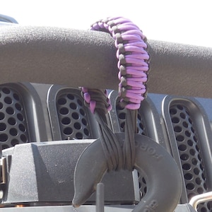 May include: A black and purple paracord grab handle is attached to a black metal hook. The handle is wrapped around a black bar. The hook is attached to a vehicle.