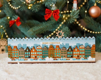 Op de afbeelding: Houten adventskalender met een stadsgezichtontwerp. De huizen zijn in tinten oranje, bruin en blauw. De kalender heeft de nummers 1-24 en de tekst "MERRY CHRISTMAS". Er valt sneeuw.
