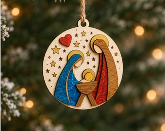 May include: A circular wooden ornament featuring a layered nativity scene with a blue-robed figure, a red-robed figure, and a central infant in a manger. The design includes a red heart and gold stars, suspended by a twine loop for hanging.