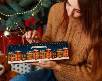 Op de afbeelding: Een houten adventskalender met een rij kleurrijke huizen onder een sterrenhemel. De kalender heeft 24 genummerde deuren en de woorden "MERRY CHRISTMAS" worden weergegeven. De kalender wordt vastgehouden door een persoon die een bruine trui en een blauwe spijkerbroek draagt.