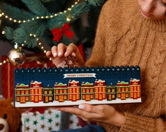 Op de afbeelding: Een houten kerst adventskalender met een rij kleurrijke huizen onder een sterrenhemel. De kalender heeft de nummers 1-24 en de woorden "MERRY CHRISTMAS". De huizen zijn rood, groen en geel.
