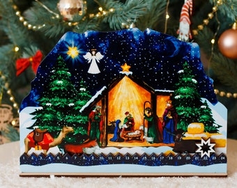 Op de afbeelding: Een houten adventskalender met de kerststal. De kalender toont een sterrenhemel, een engel en een stal met de Heilige Familie. De cijfers 1-24 staan onderaan. De kalender staat tegen een kerstboom.
