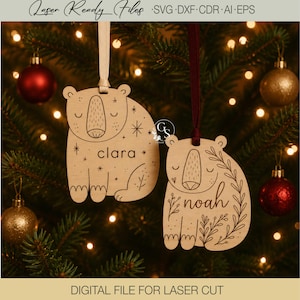 Peut inclure: Deux décorations en bois clair en forme d'ours avec des détails gravés au laser. Un ours porte le prénom Clara avec des étoiles, et l'autre le prénom Noah avec des motifs de vignes florales. Les deux sont suspendus par des rubans.