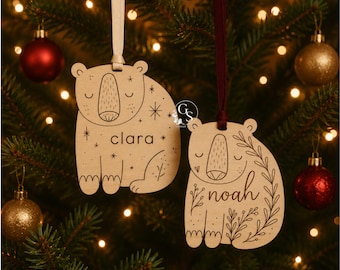 May include: Two light-colored wooden bear-shaped ornaments with laser-engraved details. One bear features the name Clara with star accents, and the other features the name Noah with floral vine accents. Both hang from ribbons.