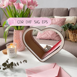 May include: A wooden heart-shaped box with a white interior, sitting on a white table with a pink envelope and a candle. The box is decorated with a wood grain pattern. The candle is labeled "Soy Wax Blend, Autumn Morning".
