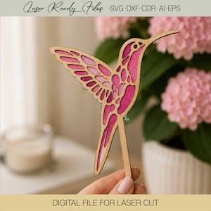 May include: A laser-cut wooden hummingbird ornament featuring intricate layered cutouts with a vibrant pink and magenta gradient finish. The bird is depicted in a mid-flight pose with a long, slender tail and a pointed beak, attached to a thin wooden stake.