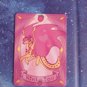 May include: A rectangular card with a cartoon dragon illustration. The dragon has pink wings and a yellow body, set against a pink background. The card includes a strawberry and the word "Kinkajou" on a banner.