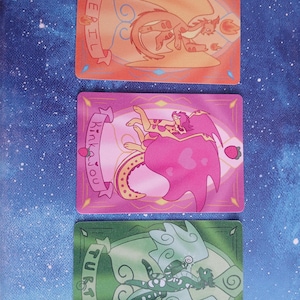 May include: Three fantasy art trading cards with dragon illustrations. The top card is orange with a dragon and the word "Peril". The middle card is pink with a dragon and the word "Kinkajou". The bottom card is green with a dragon and the word "Turtle".