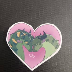 May include: A heart-shaped sticker featuring two green and yellow dragons in a loving embrace, set against a pink background. Two small hearts are drawn below the dragons. The sticker has a white border.