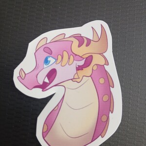 May include: A sticker depicting a cartoon dragon's head. The dragon has pink scales, a cream-coloured underbelly, and light blue eyes. Its horns and frills are a golden-yellow colour. The dragon's mouth is open, revealing sharp teeth.